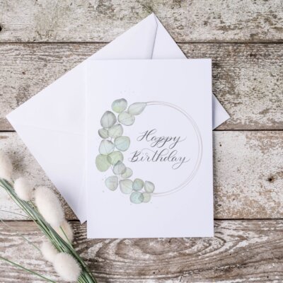Happy Birthday card Happy birthday card staged with white envelope on distressed wood with a plant shoot. Card features watercolor eucalyptus wreath with "happy birthday" written in calligraphy
