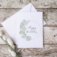 Happy birthday card staged with white envelope on distressed wood with a plant shoot. Card features watercolor eucalyptus wreath with "happy birthday" written in calligraphy